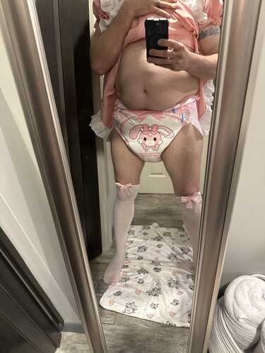 A person poses for a mirror selfie in a pink frilly outfit, diaper, and white stockings inside a bathroom.