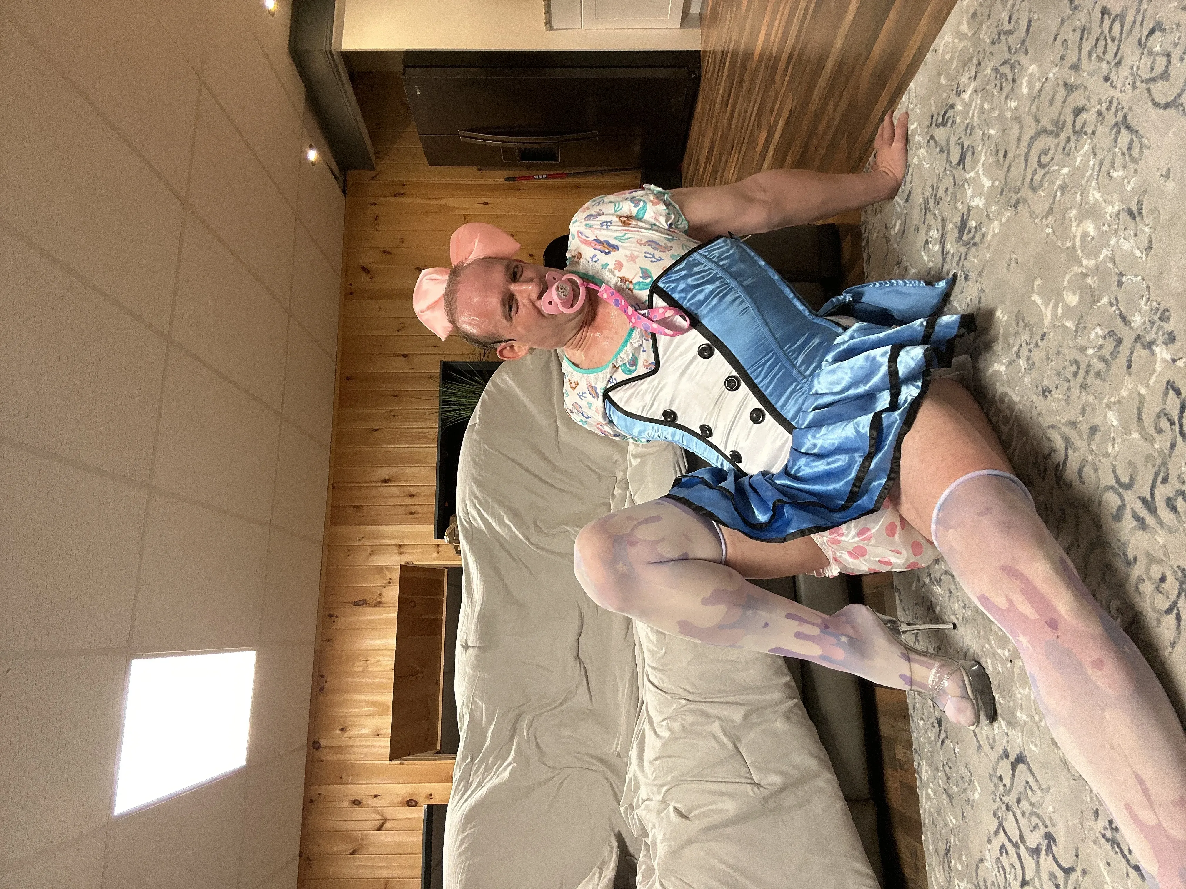 A person in a blue costume sits sideways on a sofa in a wood-paneled room with a pink bow and pacifier.