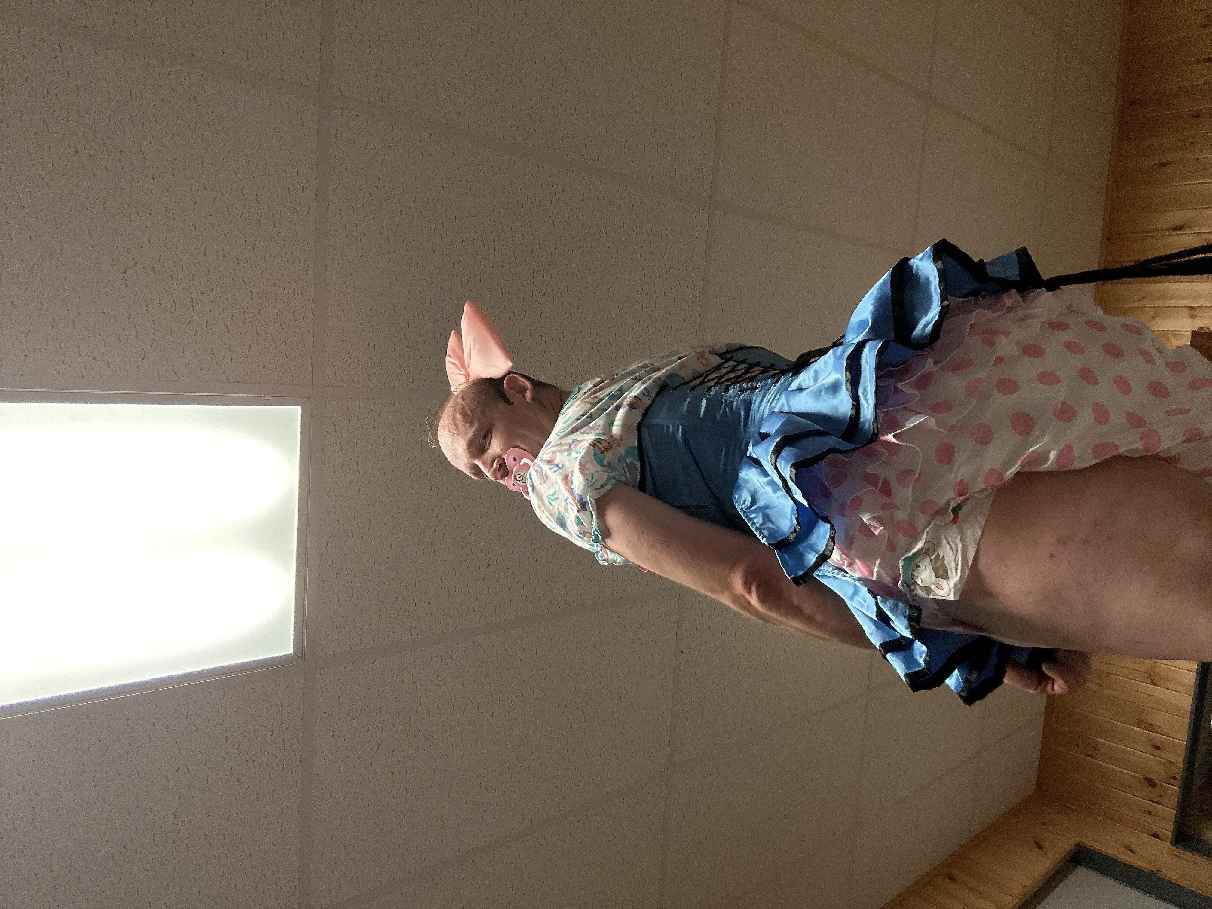 Low-angle rotated indoor portrait of a person in a blue ruffled dress and pink polka-dot outfit with a pacifier.