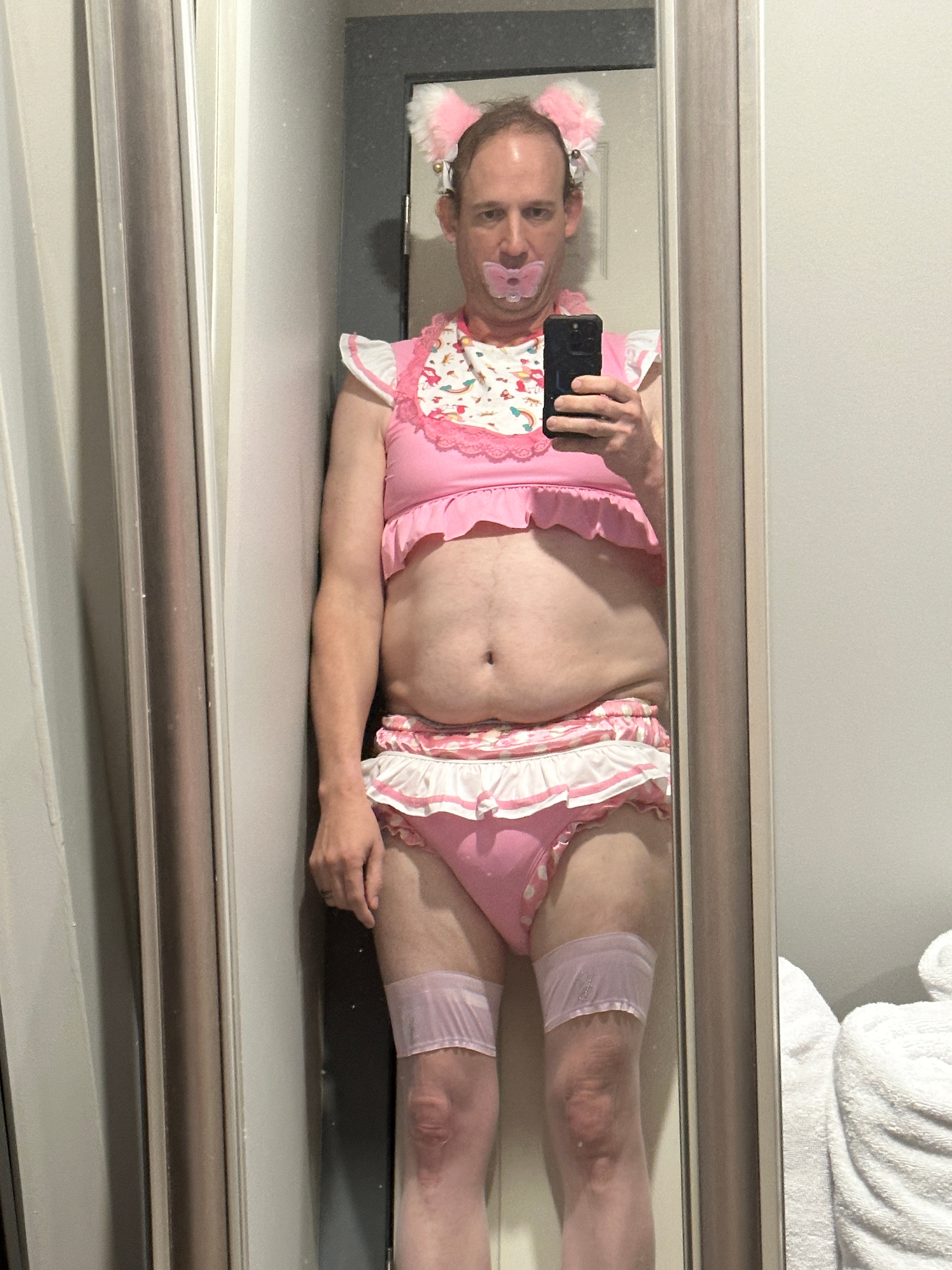 A person takes a mirror selfie in a pink frilly outfit with matching cat-ear accessories and a pink pacifier-style mouthpiece.