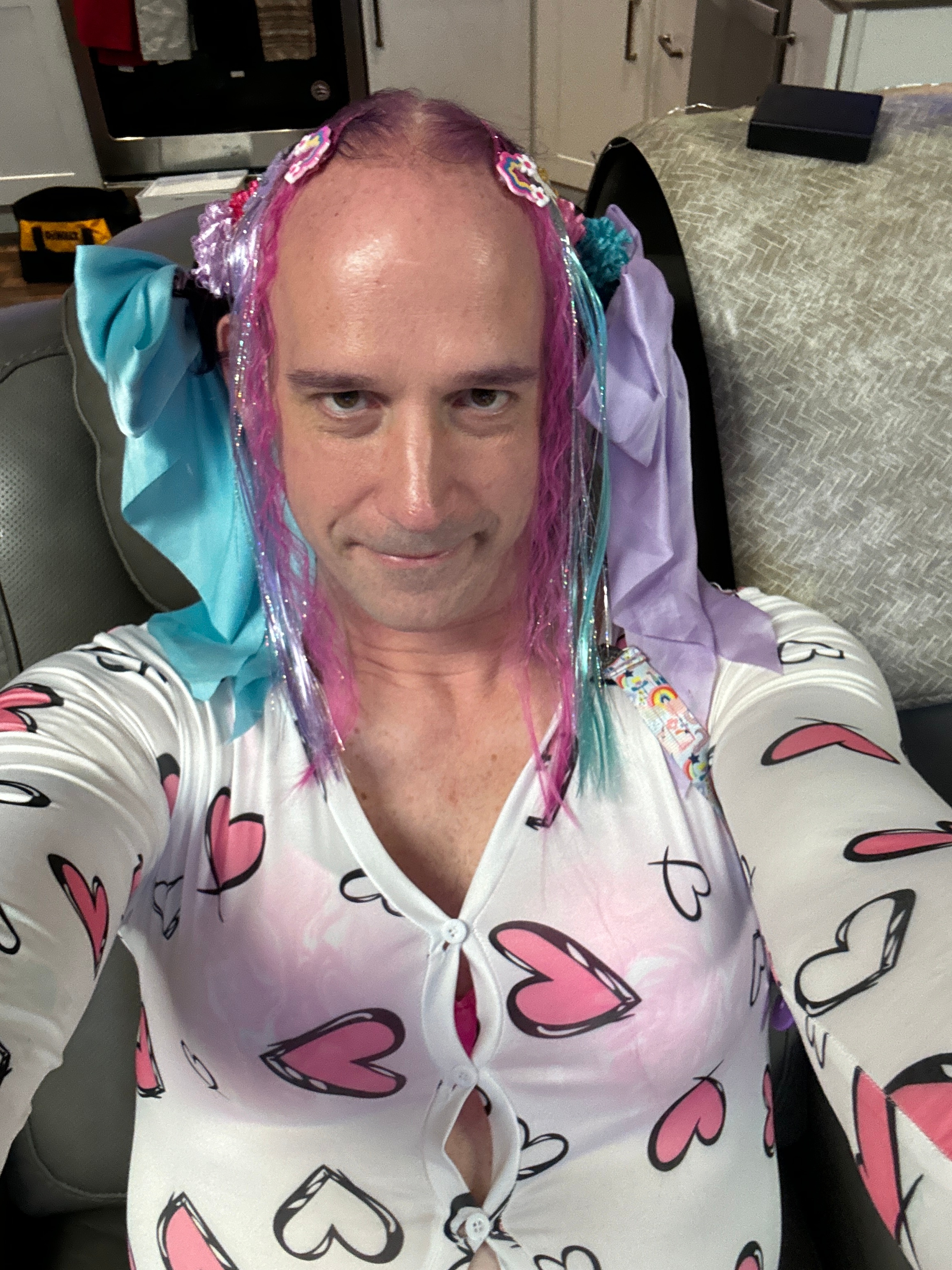 A close selfie shows a person with pink and purple hair accessories wearing a white heart-print top.