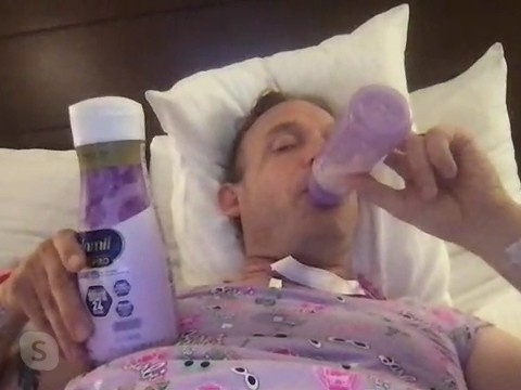A person reclines on white pillows, holding a lavender bottle and a bottle-shaped object near the mouth.
