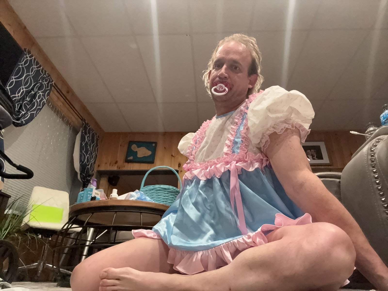A man in a blue-and-pink frilly outfit sits cross-legged on the floor with a pacifier in his mouth.