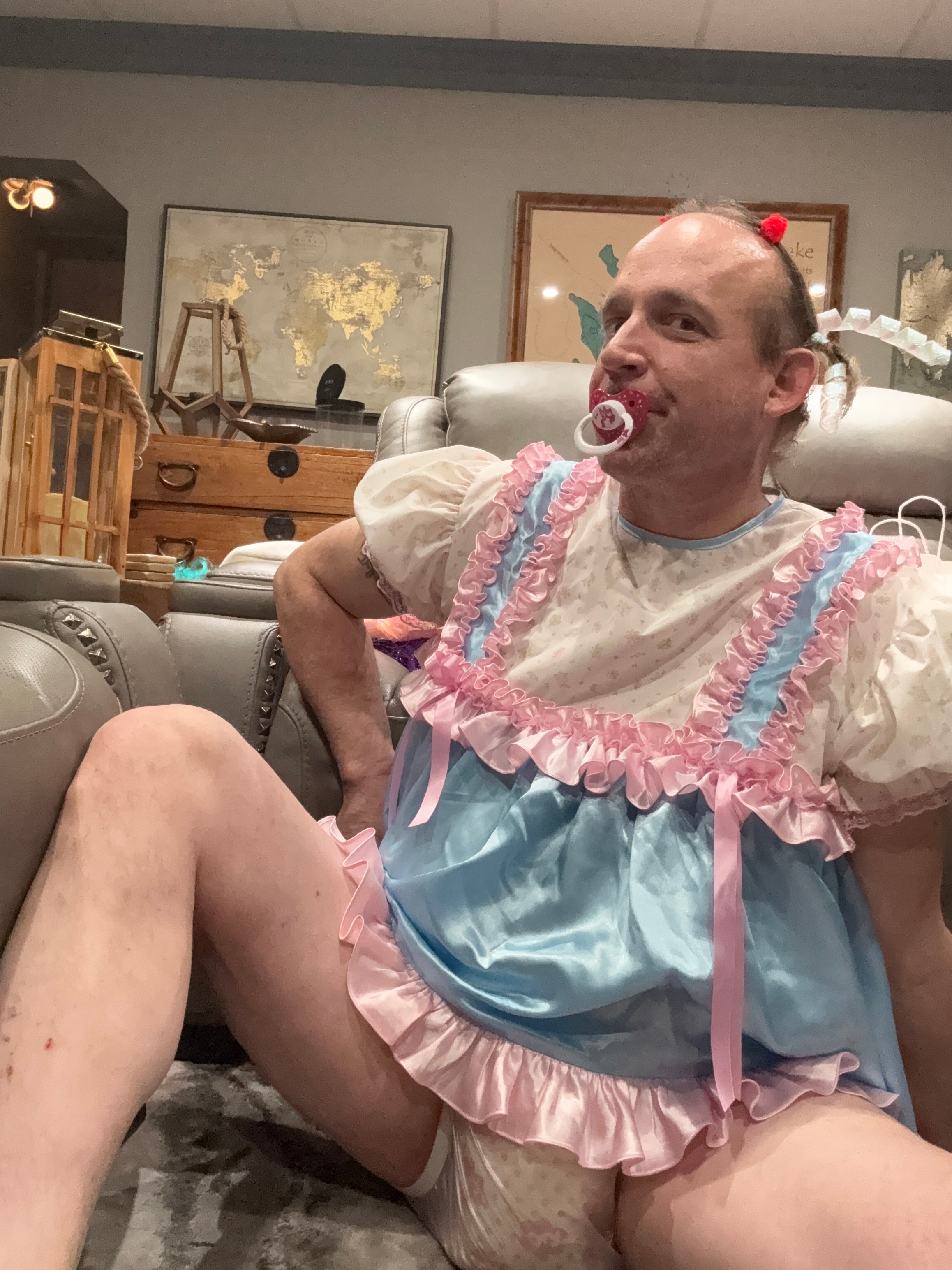 A man in a pastel ruffled outfit reclines indoors with a pacifier and one knee raised.