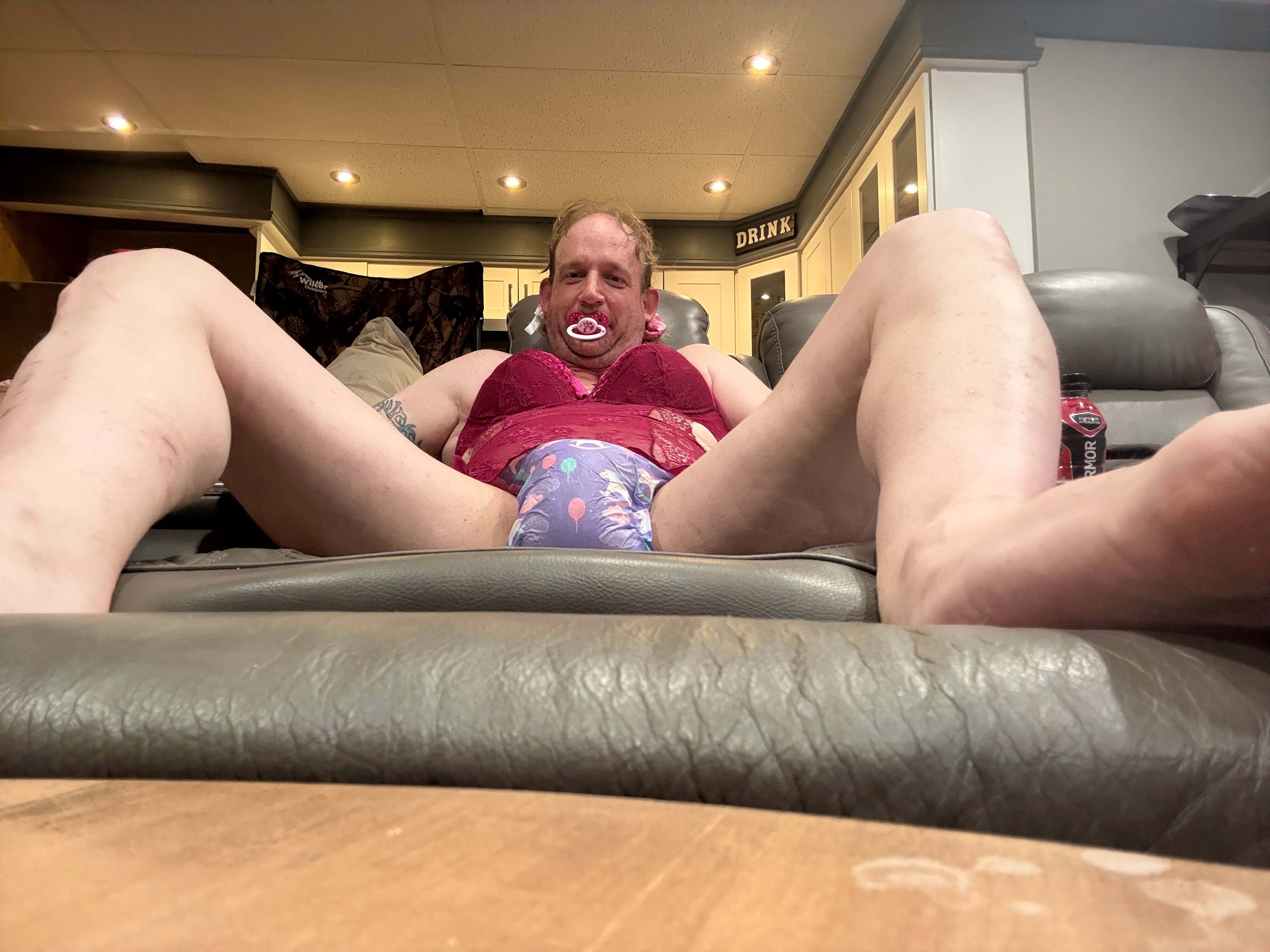 Low-angle view of a person reclining on a couch in a magenta lace top with a pacifier and legs extended toward the camera.