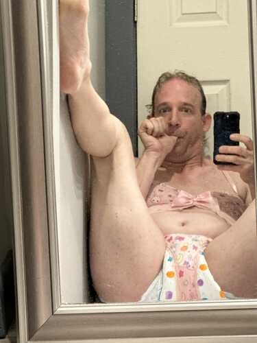 Bathroom mirror selfie of a person in a Victoria's Secret pink bra and pastel underwear posing with one leg raised.