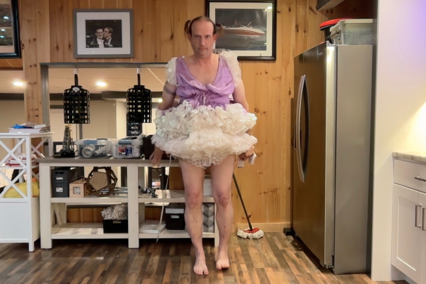 A barefoot person in a lavender top and white ruffled skirt stands in a kitchen with wood-paneled walls.