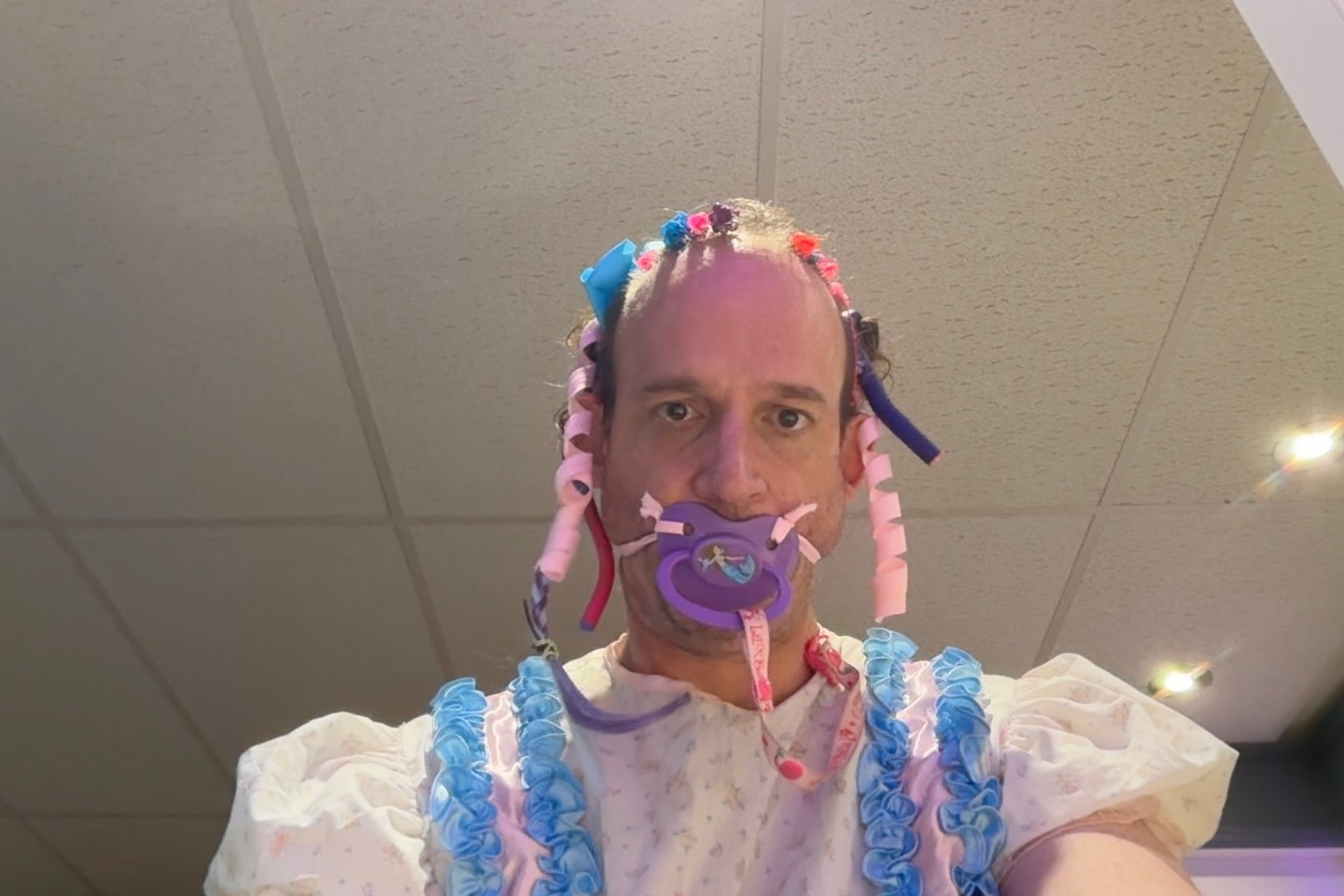 A close-up portrait shows a person with a frilly headpiece and a purple pacifier against a ceiling background.
