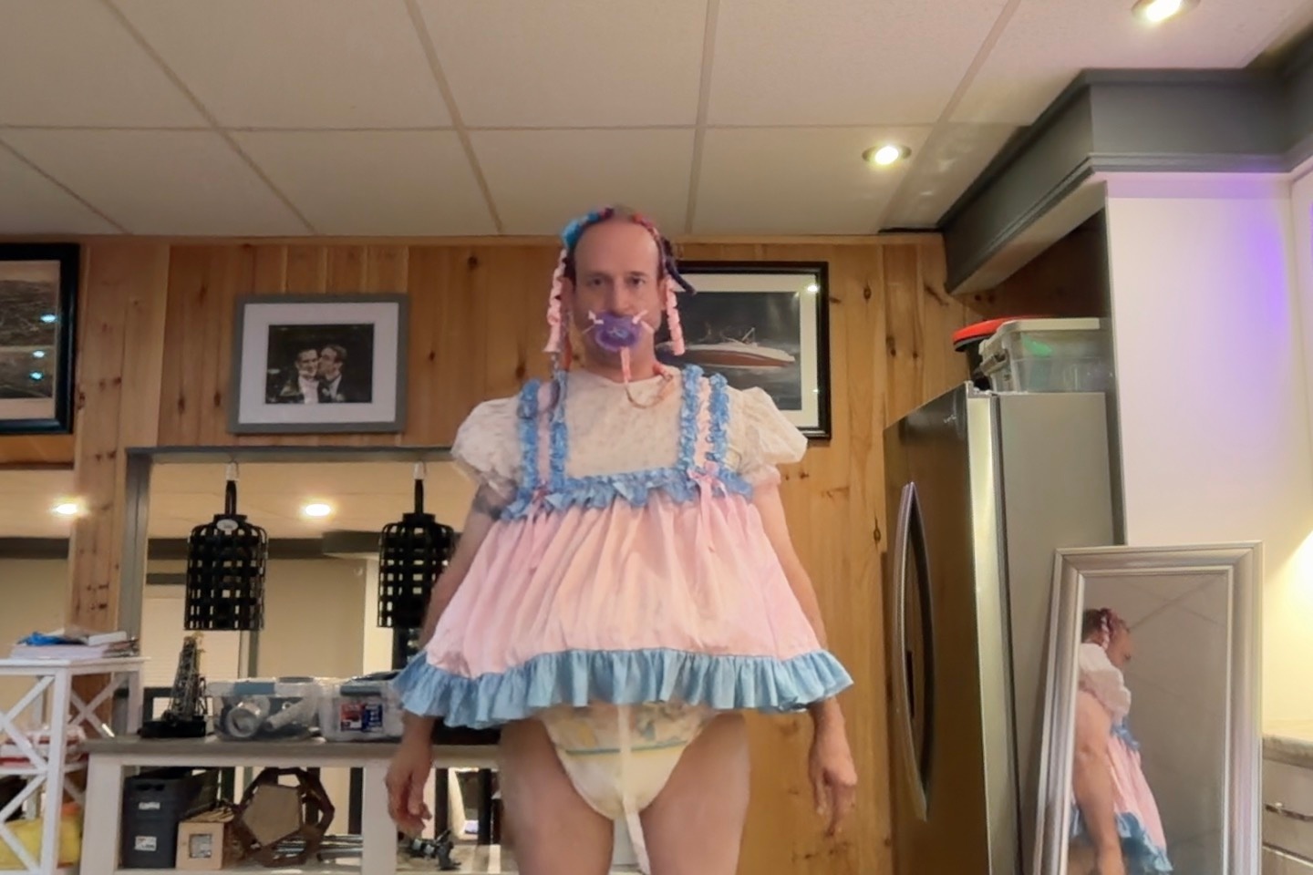 A person stands in a kitchen wearing a loose pink-and-blue frilled dress with a visible diaper underneath.