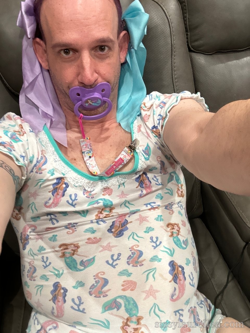 Close selfie of a person in a sea-creature print top with pastel bows and a purple pacifier, seated on a leather chair.